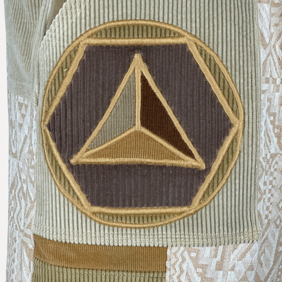 nowAwear shorts featuring a beige corduroy patch with an embroidered geometric triangle logo in brown and gold tones.