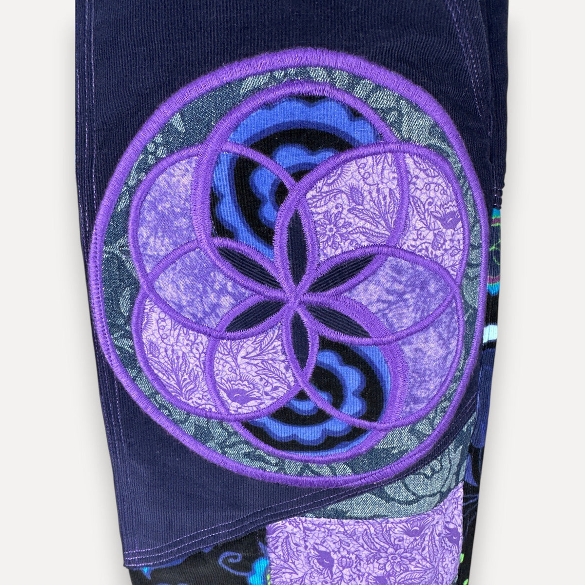 Close-up of a dark navy denim garment featuring a large circular purple and lavender floral geometric patch on the leg, with a patterned fabric patch below it, set against a plain off-white background.