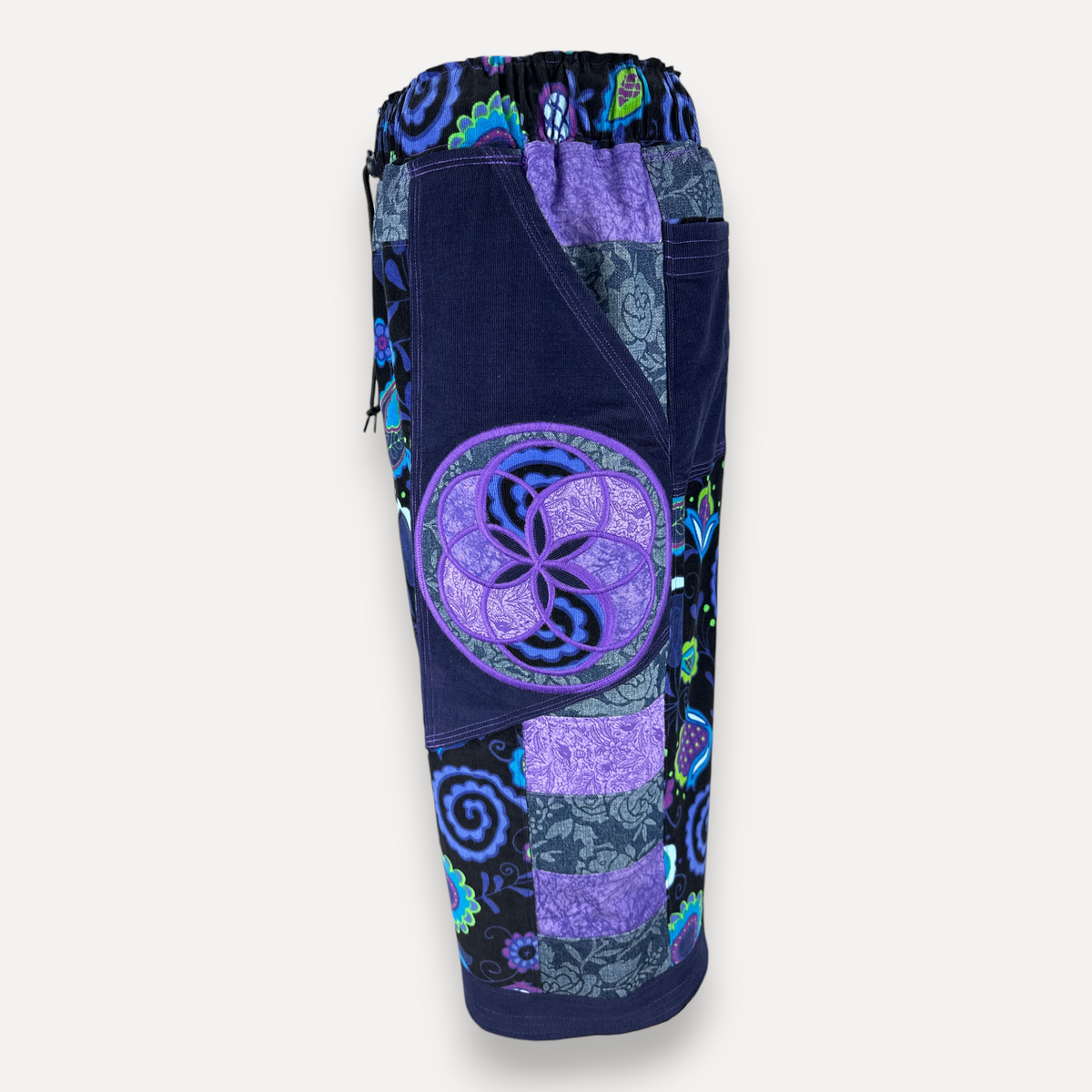 Side view of nowAwear shorts. The item has a patchwork design with dark navy, purple, blue, and teal floral/abstract patterns, plus a large circular purple emblem on the front.