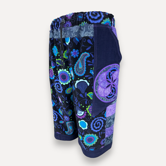 A pair of unisex patchwork shorts featuring a vibrant paisley and floral pattern in shades of blue, purple, and green on a dark navy base, with a solid navy front pocket, shown against a light gray background.